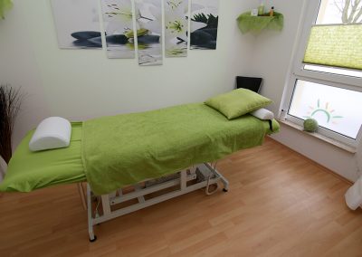 Behandlungsliege mit grünem Bezug und grünem Handtuch, dahinter ein mehrteiliges Wandbild